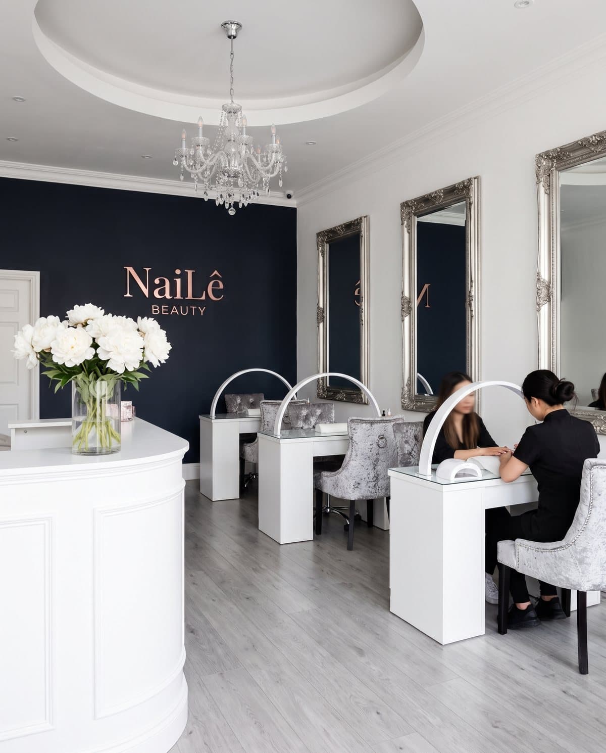 The salon: the reception desk, the sign wall, and desks under the silver mirrors with a client having her nails done